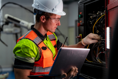 Eine Person mit Helm, Schutzbrille und Computer arbeitet an einer Schalttafel