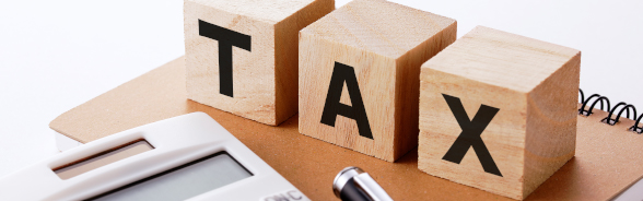 Three wooden blocks marked with the letters t, a and x are placed on a notebook. In the foreground are a calculator and a pen