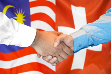 Two people shake hands against a backdrop of the Malaysian and Swiss flags