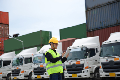 Un uomo con un gilet e un caschetto da cantiere ispeziona una zona piena di camion e container.